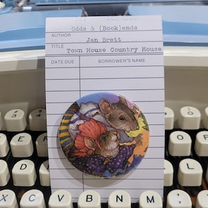 May include: A colorful button featuring an illustration of two mice from the book "Town Mouse Country Mouse" by Jan Brett. The button is placed on a library card, resting on a vintage typewriter keyboard.