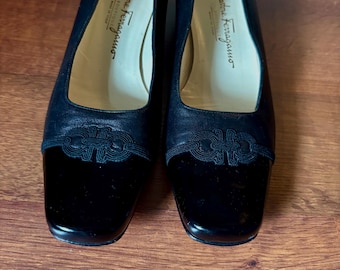 May include: A pair of black leather Salvatore Ferragamo pumps. The shoes have a square toe and a decorative design on the vamp. The heels are a medium height. The shoes are displayed on a wooden surface.