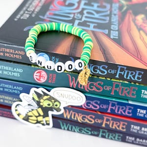 May include: A green and yellow beaded bracelet spelling "SNUDOO" with a gold charm, resting on a stack of "Wings of Fire" books. A sticker with a cartoon dragon and the word "SNUDOO" is also visible.