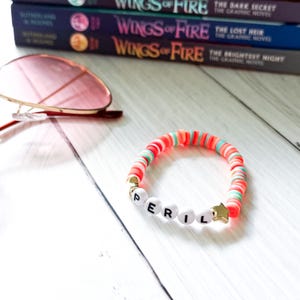 Op de afbeelding: Een kleurrijke kralenarmband met het woord "PERIL" gespeld in witte letterkralen. De armband heeft rode, roze en groene kralen en een gouden sterbedel. Zonnebril en boeken zijn op de achtergrond te zien.