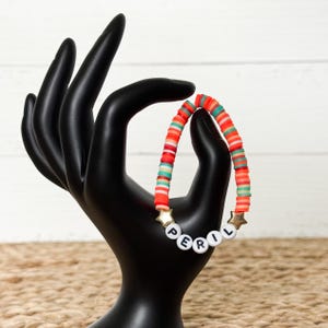 Op de afbeelding: Een kleurrijke kralenarmband met het woord "PERIL" gespeld in witte letterkralen. De armband heeft rode, oranje en turquoise schijfkralen, met gouden steraccenten. De armband wordt getoond op een zwarte hand.
