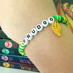Puede incluir: Una pulsera de cuentas que deletrea "SNUDOO" con cuentas de letras blancas. La pulsera presenta cuentas a rayas verdes y amarillas y un dije de hoja dorada. La pulsera se usa en la mu&ntilde;eca.