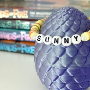 May include: A purple, egg-shaped container with a scale-like texture, adorned with a beaded bracelet spelling "SUNNY." The container sits in front of a stack of books with the title "Wings of Fire."