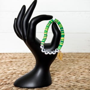 Puede incluir: Una pulsera de cuentas verdes, amarillas y blancas con la palabra "SNUDOO" escrita con cuentas de letras blancas. Un dije de hoja dorada cuelga de la pulsera. La pulsera se exhibe en un soporte de joyer&iacute;a negro en forma de mano.