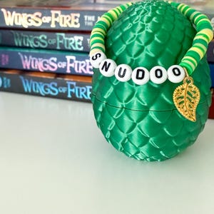 Puede incluir: Un recipiente verde, texturizado, en forma de huevo con una pulsera de cuentas que deletrea "SNUDOO". La pulsera es verde, amarilla y blanca, y tiene un dije de hoja dorada. Libros con el t&iacute;tulo "Wings of Fire" est&aacute;n en el fondo.