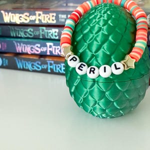 Op de afbeelding: Een groene, eivormige container met een getextureerd oppervlak, versierd met een kleurrijke kralenarmband met de tekst "PERIL". De armband heeft gouden steraccenten. De achtergrond bevat verschillende boeken met de titel "Wings of Fire".
