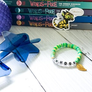 Puede incluir: Una pulsera colorida con cuentas verdes, amarillas y blancas que deletrean "SNUDOO" y un dije de hoja dorada. La pulsera se muestra con una pila de libros "Wings of Fire" y un accesorio azul en forma de estrella.
