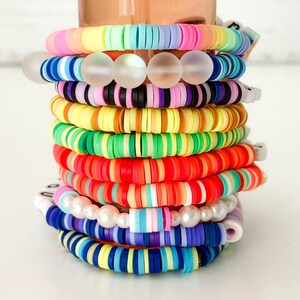 Puede incluir: Una pila de pulseras coloridas. Las pulseras est&aacute;n hechas de cuentas planas y redondas en varios colores, incluyendo rosa, amarillo, azul, verde y naranja. Algunas pulseras tienen cuentas redondas transparentes o perlas blancas. Las pulseras est&aacute;n apiladas.
