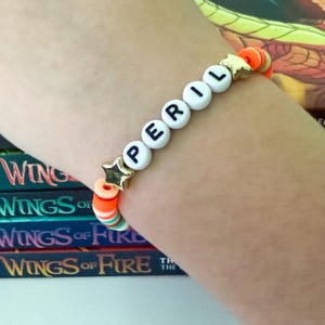 Op de afbeelding: Een kralenarmband met de tekst "PERIL" in zwarte letters op witte kralen. De armband heeft gouden ster-vormige accenten en kleurrijke oranje, groene en witte kralen. De armband zit om een pols, met de boekenreeks "Wings of Fire" op de achtergrond.