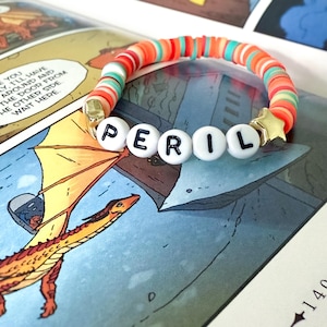 Op de afbeelding: Een kleurrijke armband met het woord "PERIL" gespeld met witte letterkralen. De armband heeft oranje, rode en groene kralen en een gouden sterbedel. De armband ligt op een stripboekpagina.