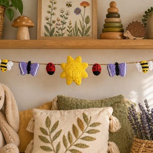 Può includere: Una ghirlanda decorativa con farfalle, api, coccinelle e un sole giallo all'uncinetto. La ghirlanda è infilata su uno spago naturale ed è esposta sopra un coniglietto di peluche e cuscini decorativi. La tavolozza dei colori comprende sfumature di verde, giallo, viola, rosso e nero.