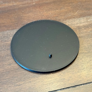 May include: A black, circular stand with a textured surface and a small, cylindrical peg. The stand is made of a solid material and is designed to hold an object. The stand is on a wooden surface.