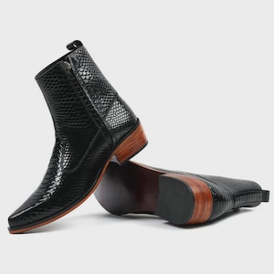 Könnte beinhalten: Schwarze Leder-Ankle Boots mit Schlangenleder-Textur. Die Stiefel haben eine spitze Zehenpartie, einen seitlichen Reißverschluss und einen Holzabsatz. Ein Stiefel steht, der andere liegt.