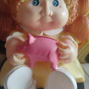 Op de afbeelding: Een vintage Cabbage Patch Kids beeldje met een roze spaarvarken. De pop heeft blauwe ogen, blond krullend haar en draagt een gele jurk met witte schoenen. Het spaarvarken is effen roze.