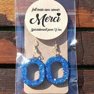 Peut inclure: Une paire de boucles d'oreilles en résine pailletée bleue, de forme ovale carrée, suspendues à des crochets dorés. Les boucles d'oreilles sont présentées sur une carte avec le texte "fait main avec amour Merci Spécialement pour Vous."