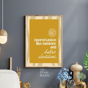 Pode incluir: Uma impressão emoldurada com fundo amarelo mostarda e o texto "Aperol tastes like summer and better decisions." Um gráfico de fatia de cítricos está no topo. A moldura é de cor de madeira clara.