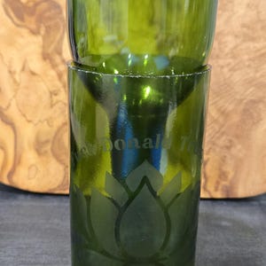 May include: Green glass drinking glass with a lotus flower design etched on the front. The glass has the words "Donald T" etched on the front. The glass is made from a recycled wine bottle.