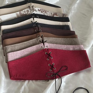 Peut inclure: Une collection de ceintures larges style corset dans diverses couleurs et textures, incluant noir, beige, marron, rose et rouge. Chaque ceinture présente une fermeture à lacets frontale avec des œillets métalliques et des cordons fins.
