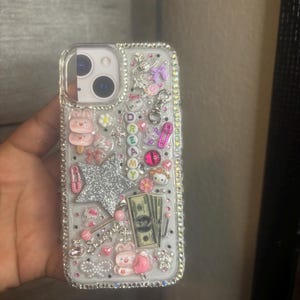 May include: Clear phone case decorated with various charms, including a silver glitter star, pink bows, and a Hello Kitty figure. The word "DREAMY" is spelled out with colorful letters. The case is embellished with rhinestones and a small stack of dollar bills.