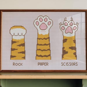 May include: A framed cross-stitch artwork depicting cat paws in a game of Rock, Paper, Scissors. The artwork features three cat paws with the words "Rock", "Paper", and "Scissors" below each paw. The paws are tan and white with pink paw pads. The frame is brown.