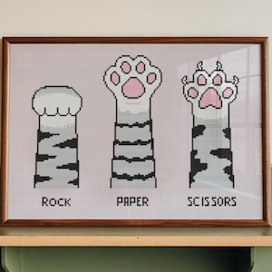 May include: Framed artwork featuring a cross-stitch design of cat paws representing Rock, Paper, and Scissors. The paws are white with pink pads and gray striped legs, set against a light pink background. The artwork is displayed on a green and wood-toned shelf.