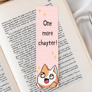 Peut inclure: Un livre ouvert avec un marque-page rose avec l'inscription "One more chapter!" et un dessin de corgi. Une tasse de café, des boules décoratives, un stylo et un pull sont également dans l'image.