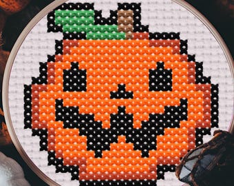May include: A cross-stitch embroidery of a jack-o'-lantern with a smiling face, green leaf, and brown stem. The pumpkin is orange with black details, set in a wooden embroidery hoop. The background includes pumpkins and fall foliage.