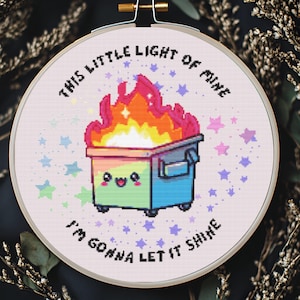 Può includere: Un telaio da ricamo a punto croce con un colorato cassonetto dei rifiuti in fiamme e la scritta "THIS LITTLE LIGHT OF MINE I'M GONNA LET IT SHINE". Il cassonetto ha una faccia sorridente ed è circondato da stelle colorate.