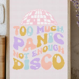 Puede incluir: Una impresión enmarcada con el texto "TOO MUCH PANIC NOT ENOUGH DISCO" en letras coloridas de estilo retro. La impresión presenta un gráfico de bola de discoteca y se muestra en una estantería de madera con libros y un jarrón de lavanda.