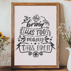 Puede incluir: Un cuadro enmarcado con el texto "My brain has too many tabs open" en escritura negra. El cuadro está sobre una mesa de madera con una cesta de flores amarillas y una jarra blanca con flores blancas.