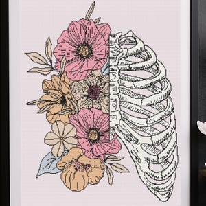 May include: A framed art print featuring a floral and anatomical design. The artwork is split vertically, with a rib cage on the right and a collection of pink, yellow, and peach flowers on the left. The print is in a black frame.