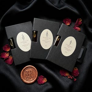 May include: Three black boxes with oval labels and small glass vials. The labels read "Ghost of the Temple", "Blood of the Lotus", and "Starwoven Veil". A bronze wax seal and scattered rose petals are also visible.
