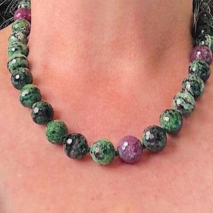 May include: A beaded necklace featuring large, round beads in shades of green and purple. The beads have a speckled appearance, creating a unique texture. The necklace is displayed against a skin-toned background.