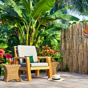 May include: A wooden outdoor chair with a light gray cushion and a small green pillow sits on a patio. A woven side table, a black planter with red flowers, and a straw hat are nearby. Lush green plants and a bamboo fence are in the background.