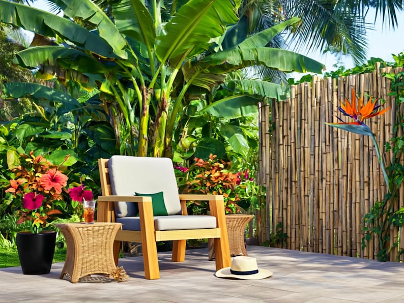 May include: A wooden outdoor chair with a light gray cushion and a small green pillow sits on a patio. A woven side table, a black planter with red flowers, and a straw hat are nearby. Lush green plants and a bamboo fence are in the background.