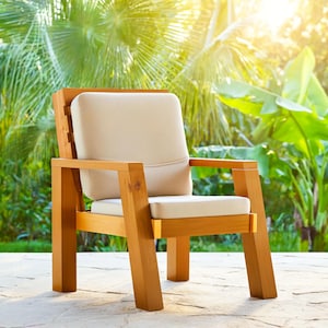 May include: A wooden outdoor armchair with a light beige cushion and backrest. The chair has a natural wood finish and is set on a stone patio. The background features lush green foliage and sunlight.