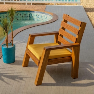 May include: A wooden outdoor chair with a yellow cushion. The chair has a slatted back and armrests. It is placed on a light gray tiled surface next to a swimming pool. A blue planter with plants is nearby.