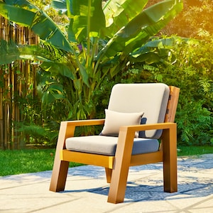 May include: A wooden outdoor armchair with a light gray cushion and a small lumbar pillow. The chair has a natural wood finish and is set on a stone patio with lush greenery in the background. The chair is designed for outdoor use.