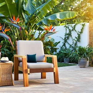 May include: Outdoor patio scene featuring a wooden armchair with a cream-colored cushion and a small, dark blue pillow. A woven side table holds a hat and a drink. Lush greenery and bird of paradise flowers surround the seating area.