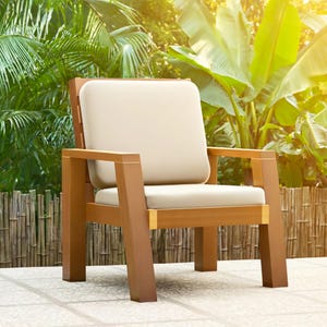 May include: A wooden outdoor armchair with a light beige cushion and backrest. The chair has a natural wood finish and is set against a backdrop of lush green foliage and a bamboo fence. The chair's dimensions are approximately 30 inches wide, 30 inches deep, and 30 inches high.