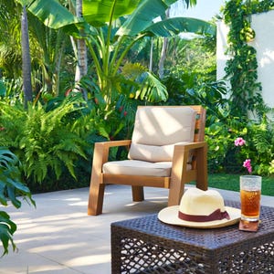 May include: A wooden outdoor chair with a beige cushion sits on a patio. A woven brown table holds a straw hat with a burgundy band, a glass of iced tea, and a coaster. Lush green plants and flowers surround the patio.