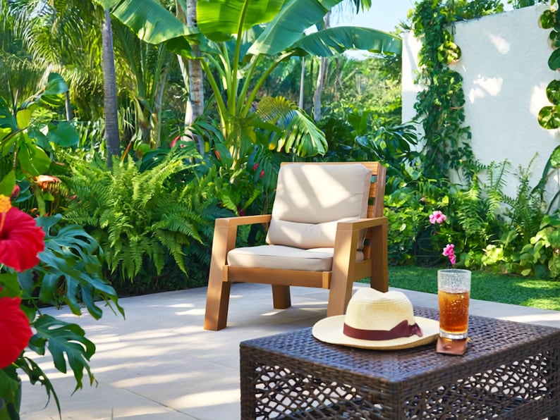 May include: A wooden outdoor chair with a beige cushion sits on a patio. A woven brown table holds a straw hat with a burgundy band, a glass of iced tea, and a coaster. Lush green plants and flowers surround the patio.