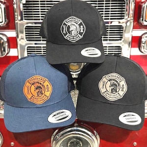 May include: Three baseball caps with mesh backs and snapback closures. One cap is blue with a tan patch, while two are black with white or tan patches. Each patch features a circular emblem with a helmet, ladders, and the text Vanguard Engine Company West Islip Fire Department.