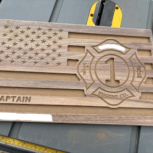 May include: Wooden plaque with an engraved American flag design. Features the text "CAPTAIN" and a fire department emblem with the number "1" and "ENGINE CO." in a circular design. The plaque is made of light and dark brown wood.