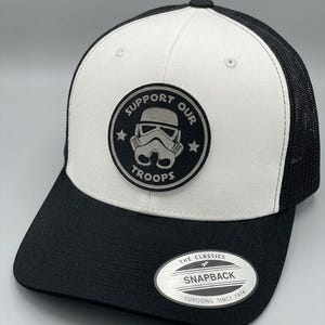 May include: A white and black snapback cap featuring a circular patch on the front panel. The patch displays a stormtrooper helmet graphic with the text SUPPORT OUR TROOPS. The cap has a black brim and a black mesh back. A sticker on the brim reads THE CLASSICS SNAPBACK YUPOONG SINCE 1974.