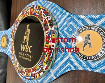 May include: A custom Muay Thai championship belt with a blue patterned strap. The belt features a large central emblem with the WBC logo and a smaller World Boxing Council emblem. The central emblem is surrounded by flags.