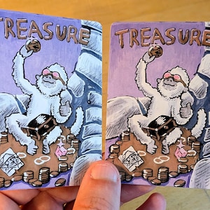 May include: Two rectangular cards featuring a cartoon illustration of a white yeti wearing pink goggles, holding a gold coin, and sitting on a pile of treasure. The word TREASURE is printed at the top in brown letters against a purple background.