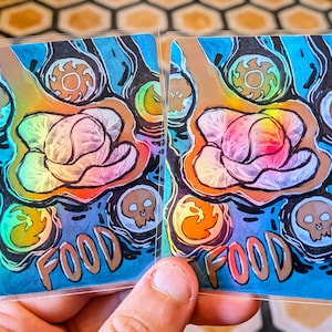 May include: Two holographic cards with a vibrant illustration. The design includes a central cabbage-like form with rainbow colours, surrounded by stylized flames, skulls, and circular patterns. The word "FOOD" is written at the bottom.