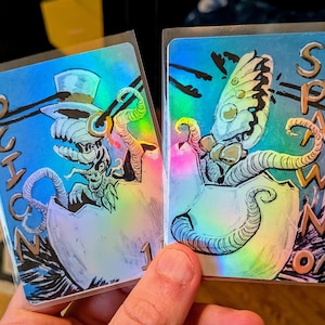 Peut inclure: Deux cartes à collectionner holographiques avec des illustrations surréalistes. L'une représente une créature en haut-de-forme sortant d'un œuf, avec le mot "SCHOON" au-dessus. L'autre montre une créature similaire avec des ailes de papillon, avec le mot "SPAWN" au-dessus.