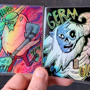 May include: Two illustrated cards featuring fantasy characters. One card reads "Kaldra" with a green-skinned figure and a long beard. The other card says "Germ" with a blue-faced creature. Both cards have colorful backgrounds.